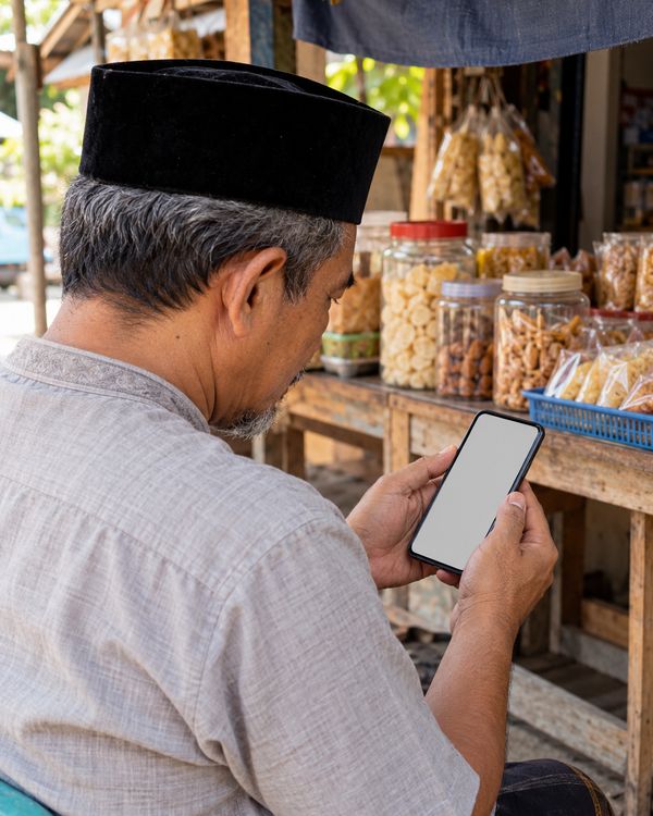 Foto suasana (dibuat AI, bukan foto usaha sungguhan): bapak pemilik warung camilan berpeci melihat ponselnya di depan dagangannya.