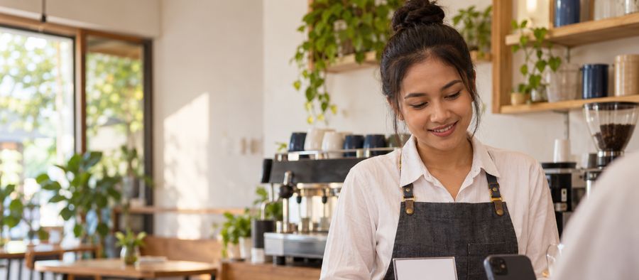Foto suasana (dibuat AI, bukan foto usaha sungguhan): pemilik kafe tersenyum di balik meja kasir sementara pembeli memegang ponsel.